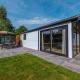 Moderne lodge Velthorst med store vinduer, fliseterrasse og havemøbler omgivet af grønne træer.