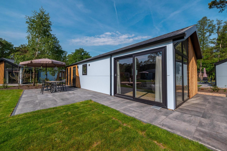 Lodge Velthorst moderno con grandes ventanas, terraza de baldosas y muebles de exterior entre árboles.