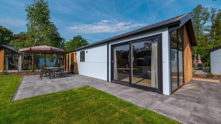 Lodge Velthorst moderne avec grandes baies vitrées, terrasse carrelée et mobilier de jardin, entouré d’arbres.