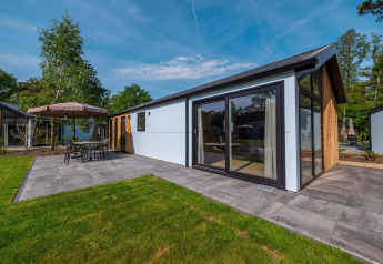 Lodge Velthorst moderne avec grandes baies vitrées, terrasse carrelée et mobilier de jardin, entouré d’arbres.