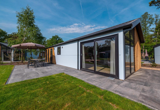 Lodge Velthorst moderne avec grandes baies vitrées, terrasse carrelée et mobilier de jardin, entouré d’arbres.