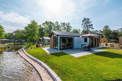 Moderne Lodge mit Terrasse und Sonnenschirm in De Hooge Veluwe, Niederlande, neben einem Teich gelegen.