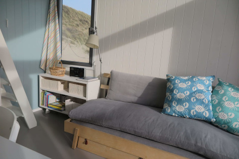Interior luminoso de casa de playa con sofá gris, cojines azules de cangrejo y ventana hacia dunas.