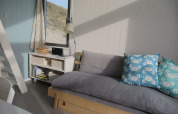 Interior luminoso de casa de playa con sofá gris, cojines azules de cangrejo y ventana hacia dunas.