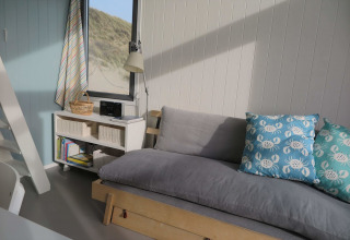 Lyst og hyggeligt strandhus interiør med grå sofa, blå puder med krabbemotiv og udsigt til klitter.