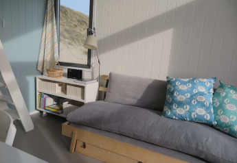 Licht en knus strandhuisinterieur met grijze bank, blauwe krabkussens en raam met uitzicht op de duinen.