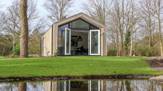 Forest Lodge con grandi porte di vetro aperte sul prato verde, circondata da alberi e un laghetto davanti.