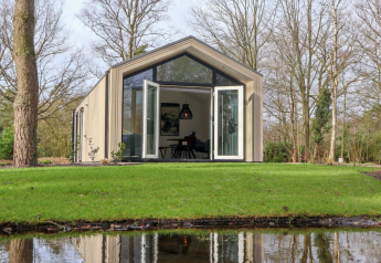 Forest Lodge mit großen Glastüren zur Wiese hin, umgeben von Bäumen und einem Teich im Vordergrund.