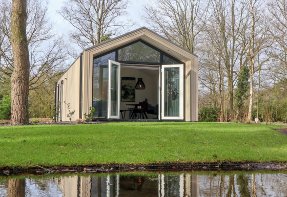 Forest Lodge con grandi porte di vetro aperte sul prato verde, circondata da alberi e un laghetto davanti.