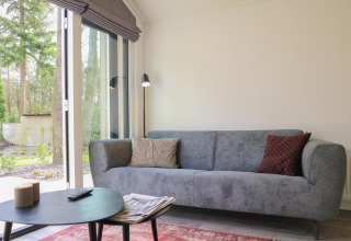 Lys stue i Forest Lodge på De Hooge Veluwe, Holland, med grå sofa og store vinduer til skovudsigt.