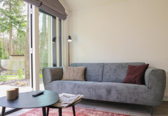 Sala luminosa en Forest Lodge, De Hooge Veluwe, Países Bajos, con sofá gris y vistas al bosque.