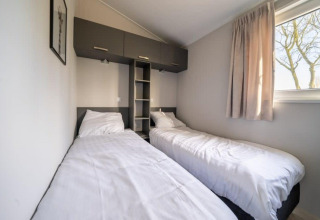 Lodge slaapkamer met twee aparte bedden, wit beddengoed, raam met gordijn en opbergruimte erboven.