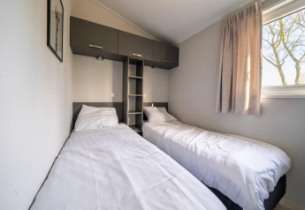 Chambre de lodge avec deux lits simples, literie blanche, fenêtre avec rideaux et placards en hauteur.