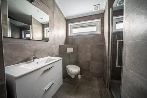 Salle de bain moderne dans un lodge avec carrelage gris, toilettes suspendues, grand miroir et douche séparée.