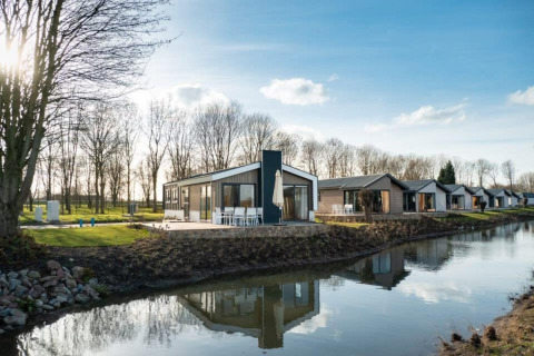 Modern lodge by a small canal, with similar cabins and leafless trees in the background on a sunny day.