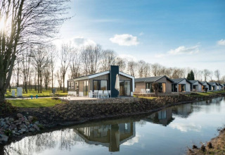 Lodge moderne au bord d’un canal, avec d’autres chalets similaires et arbres nus sous un ciel ensoleillé.