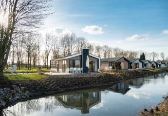Lodge moderne au bord d’un canal, avec d’autres chalets similaires et arbres nus sous un ciel ensoleillé.
