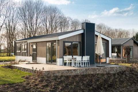 Moderne lodge met grote ramen, buitenterras en eethoek, omgeven door bomen en een natuurlijk landschap.