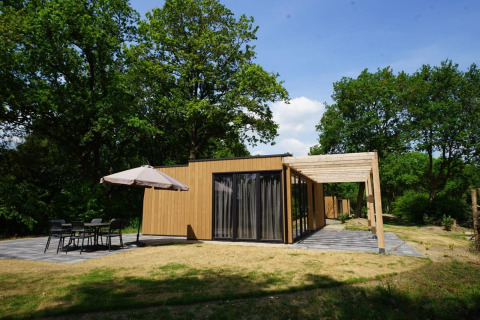 Moderne lodge ved navn Cube med træfacade, stor terrasse og udendørs siddeplads omgivet af grønne træer.