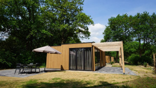 Moderne lodge ved navn Cube med træfacade, stor terrasse og udendørs siddeplads omgivet af grønne træer.