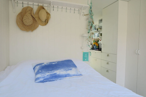 Soveværelse med hvide vægge, halmhatte på knager og et blåt pyntepude i Beach house 'Dishoek 60', Zeeland.