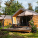 Zwei kleine Solo Retreat Holzhäuser mit Terrassen, Stühlen und Sonnenschirmen in grüner Natur.