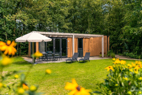 Buitenaanzicht van Cube Hot tub lodge met houten gevel, terrasmeubilair, parasol en groene tuin op de voorgrond.
