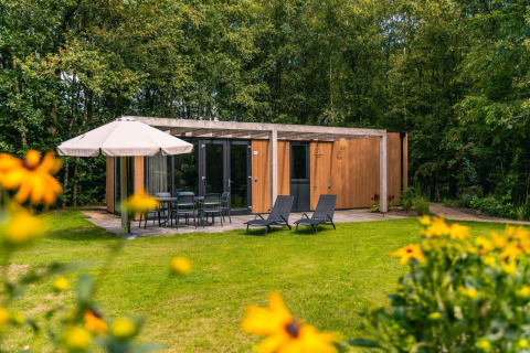 Buitenaanzicht van Cube Hot tub lodge met houten gevel, terrasmeubilair, parasol en groene tuin op de voorgrond.