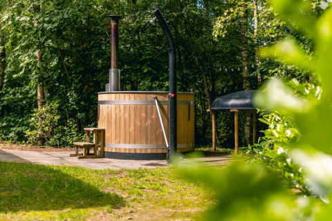 Buiten hottub van hout met schoorsteen bij Cube Hot tub lodge op EuroParcs Ruinen, Nederland, omgeven door groen.