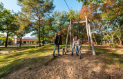 Familie mit Hund spielt an einer Seilbahn in einem sonnigen, waldreichen Glamping-Resort.