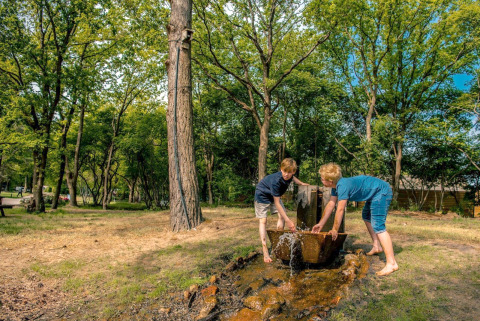 Twee kinderen spelen bij een buitenwaterbron omringd door bomen bij een glampingaccommodatie.