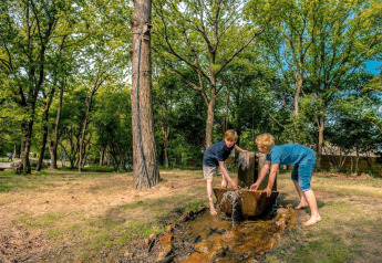 Twee kinderen spelen bij een buitenwaterbron omringd door bomen bij een glampingaccommodatie.