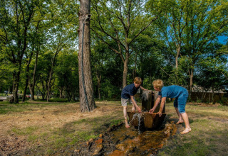 Two children play at an outdoor water source surrounded by trees at a glamping accommodation site.