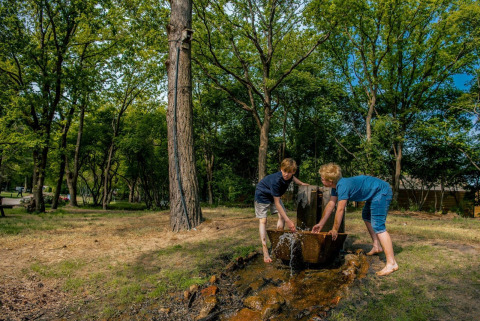 Zwei Kinder spielen an einer Wasserquelle im Freien auf einem Glampingplatz mit vielen Bäumen.