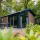 Moderne Velthorst-lodge i skovomgivelse med glasfacade, havemøbler og grønne planter i forgrunden.