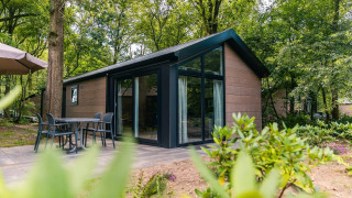 Moderne Velthorst-Lodge im Wald, mit Glasfront, Terrasse, Tisch und Stühlen, umgeben von Natur.