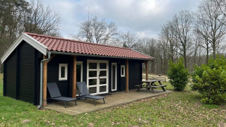 Schwarze Lodge mit rotem Dach, Holzterrasse, zwei Liegestühlen und Picknicktisch im Grünen.
