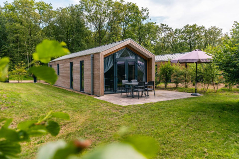 Forest Lodge eksteriør med terrasse, havemøbler og parasol, omgivet af grønne træer og græsplæne.