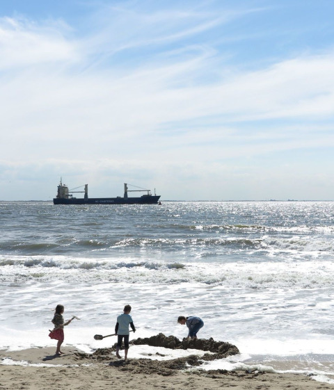 Drei Kinder spielen am Strand im Sand, während ein Frachtschiff bei Dishoek, Zeeland, vorbeifährt.