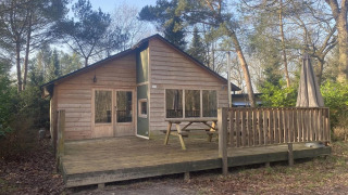 Glamping Family Lodge 10: Holzlodge mit Terrasse, Picknickbank und Sonnenschirm in bewaldeter Umgebung.