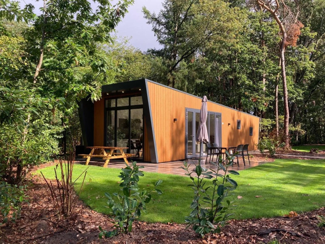 Moderne Holzhütte mit großen Fenstern im Grünen, mit Picknicktisch und Gartenmöbeln auf Terrasse.