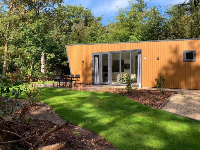 Moderne Lodge mit Holzfassade, großen Fenstern und Terrasse, umgeben von Wiese und Bäumen.