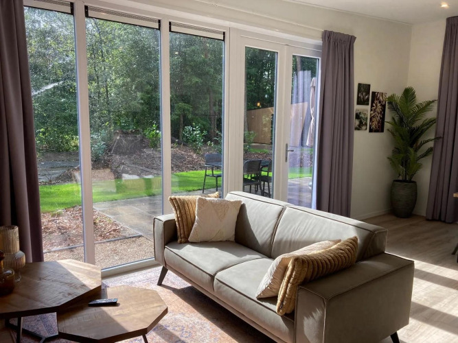 Helles Wohnzimmer in einer Lodge mit großem Sofa, bodentiefen Fenstern und Blick auf Garten und Wald.