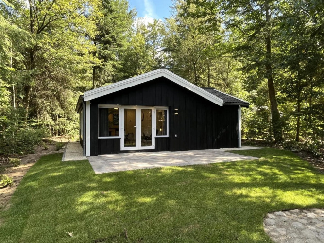 Schwarze Glamping-Holzhütte im Wald mit großer Terrasse und gepflegtem Rasen davor.