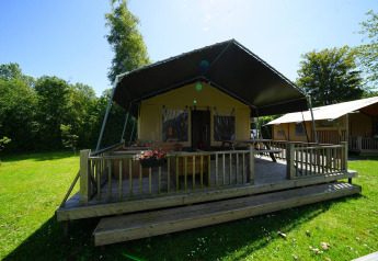 Safari-Zelt-Lodge bei Molengroet in den Niederlanden mit Veranda, grünem Rasen und Bäumen im Sonnenlicht.