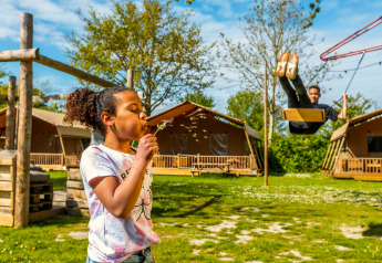 Een meisje blaast op een paardenbloem en een jongen schommelt voor lodge-tenten in de zon.
