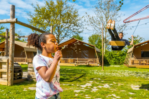 A girl blows dandelion seeds and a boy swings in front of cozy lodge tents on a sunny day.