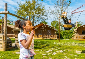 Una bambina soffia semi di tarassaco mentre un bambino dondola davanti a lodge sotto il sole.