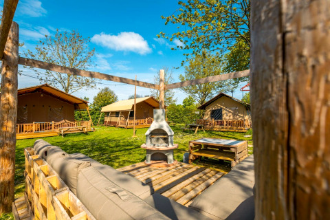 Vue extérieure d’un lodge avec des tentes, bancs en bois, pelouse verte et coin salon sous le ciel bleu.