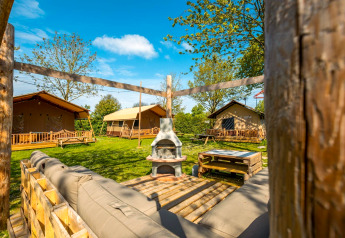 Outdoor view of a lodge with tents, wooden benches, grassy lawn, seating area, and clear blue sky above.